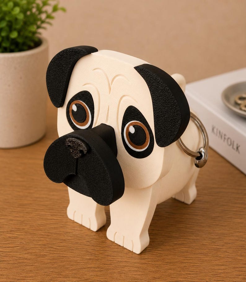 May include: A cream-colored pug figurine with black ears, nose, and accents. The dog-shaped item has large brown eyes and a metal ring attached to its side. The pug is standing on four legs and is a decorative item.