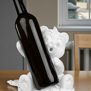 May include: A white bear-shaped wine bottle holder. The bear is holding a dark brown wine bottle. The holder is on a white, textured surface. The bear has a surprised expression.