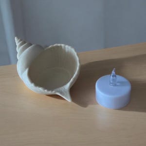 May include: A cream-colored seashell-shaped bowl with a spiral top and a small, light blue LED candle. The bowl is on a light brown surface. The candle has a clear, flame-shaped top.