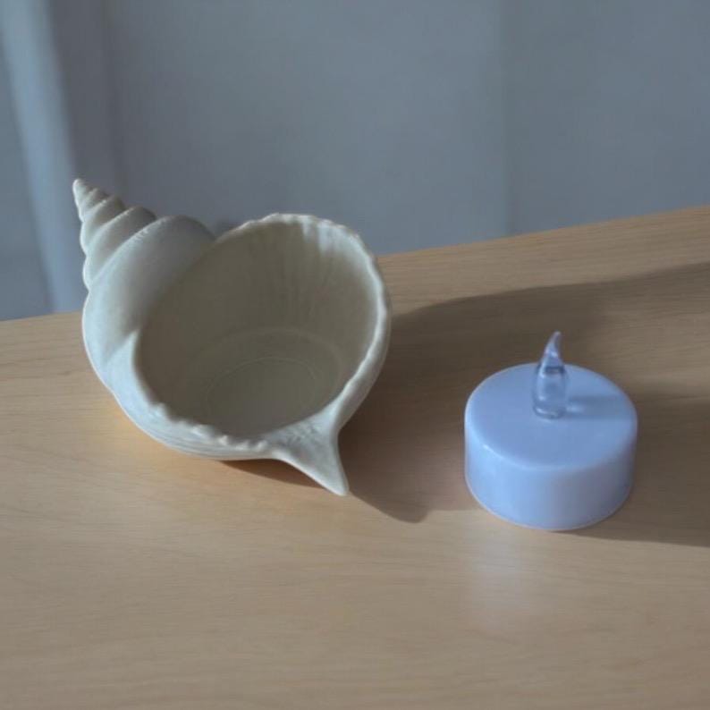 May include: A cream-colored seashell-shaped bowl with a spiral top and a small, light blue LED candle. The bowl is on a light brown surface. The candle has a clear, flame-shaped top.