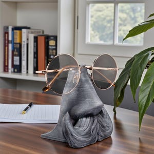May include: A gray novelty eyeglass holder shaped like a nose and mustache, holding a pair of round, gold-rimmed glasses. The holder sits on a wooden desk, with a notepad and pen nearby. A bookshelf and window are in the background.