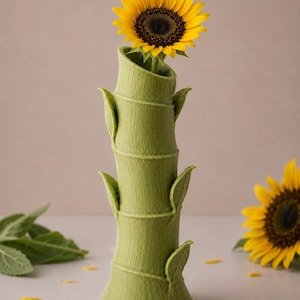Puede incluir: Un jarr&oacute;n verde con forma de bamb&uacute; sostiene un girasol amarillo vibrante. El jarr&oacute;n tiene una superficie texturizada y detalles en forma de hojas. Girasoles y p&eacute;talos adicionales est&aacute;n esparcidos alrededor del jarr&oacute;n, creando un arreglo floral natural.