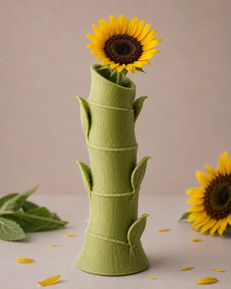 Puede incluir: Un jarr&oacute;n verde con forma de bamb&uacute; sostiene un girasol amarillo vibrante. El jarr&oacute;n tiene una superficie texturizada y detalles en forma de hojas. Girasoles y p&eacute;talos adicionales est&aacute;n esparcidos alrededor del jarr&oacute;n, creando un arreglo floral natural.