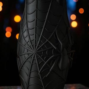 May include: Black cylindrical container with a raised spiderweb design and a spider on the side. The container has a matte finish and is set against a blurred background with orange and blue lights.