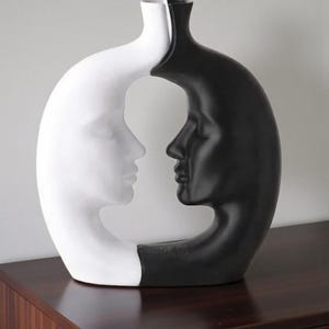 K&ouml;nnte beinhalten: Dekorative Vase mit zwei sich gegen&uuml;berliegenden menschlichen Profilen, eines wei&szlig; und eines schwarz. Die Vase hat ein einzigartiges, offenes Design, wobei sich die Profile in der Mitte treffen. Die Vase besteht aus gl&auml;nzendem Material und steht auf einer dunklen Holzoberfl&auml;che.