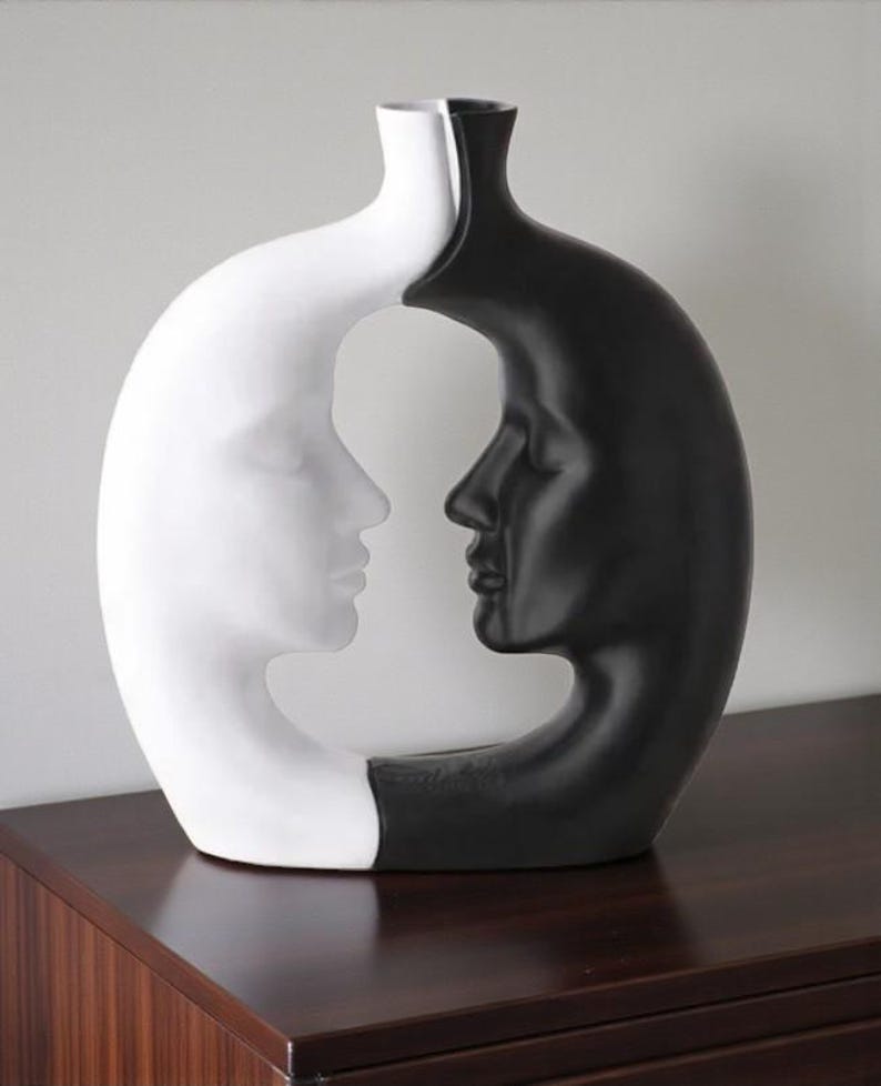 K&ouml;nnte beinhalten: Dekorative Vase mit zwei sich gegen&uuml;berliegenden menschlichen Profilen, eines wei&szlig; und eines schwarz. Die Vase hat ein einzigartiges, offenes Design, wobei sich die Profile in der Mitte treffen. Die Vase besteht aus gl&auml;nzendem Material und steht auf einer dunklen Holzoberfl&auml;che.