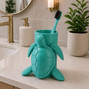 May include: A teal turtle-shaped toothbrush holder with a matching teal toothbrush. The holder is made of a solid material and has a detailed turtle design. The toothbrush has black bristles.