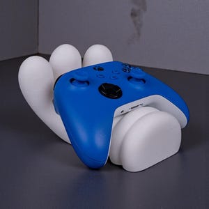 May include: A blue and white video game controller rests on a white, hand-shaped stand. The controller has a sleek design with black buttons and a white base. The stand is a modern, artistic design.