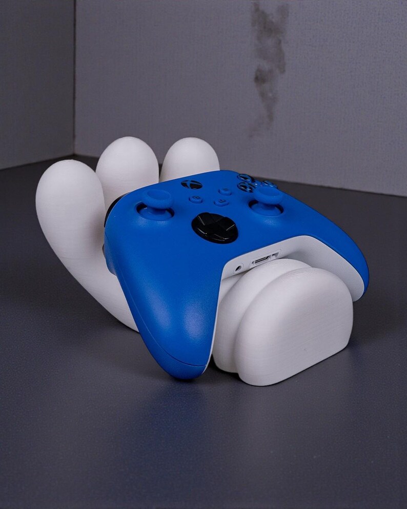 May include: A blue and white video game controller rests on a white, hand-shaped stand. The controller has a sleek design with black buttons and a white base. The stand is a modern, artistic design.
