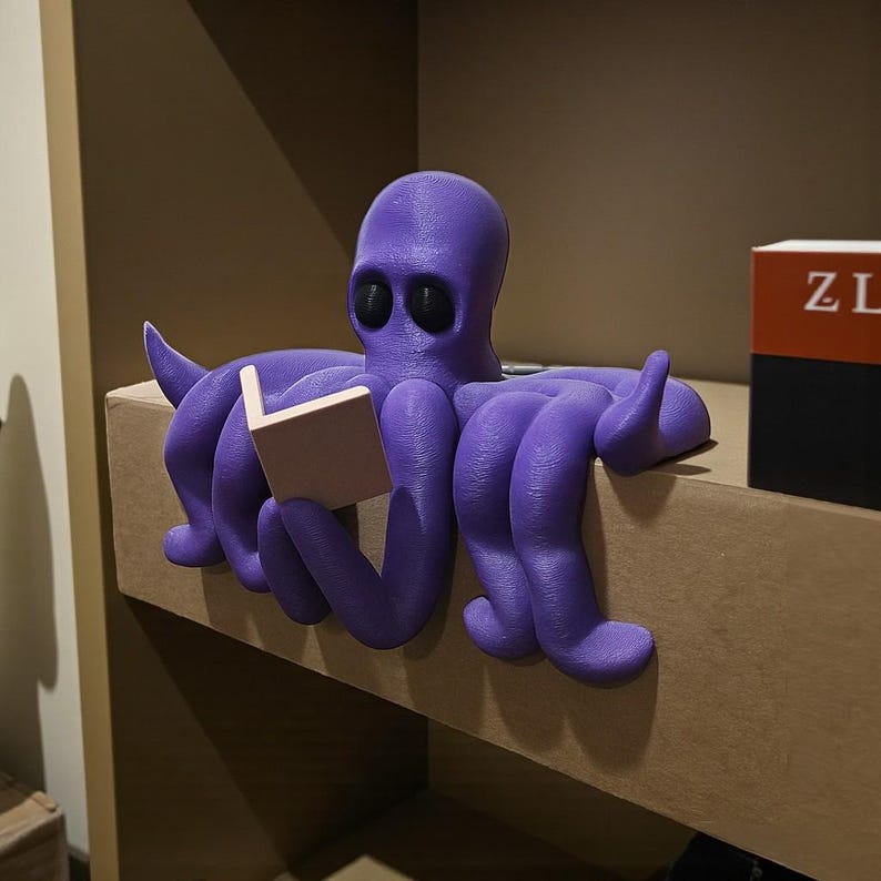 May include: A purple octopus figurine reading a small book. The octopus has large black eyes and is perched on a shelf. The book is a light tan color. The figurine is a decorative item.