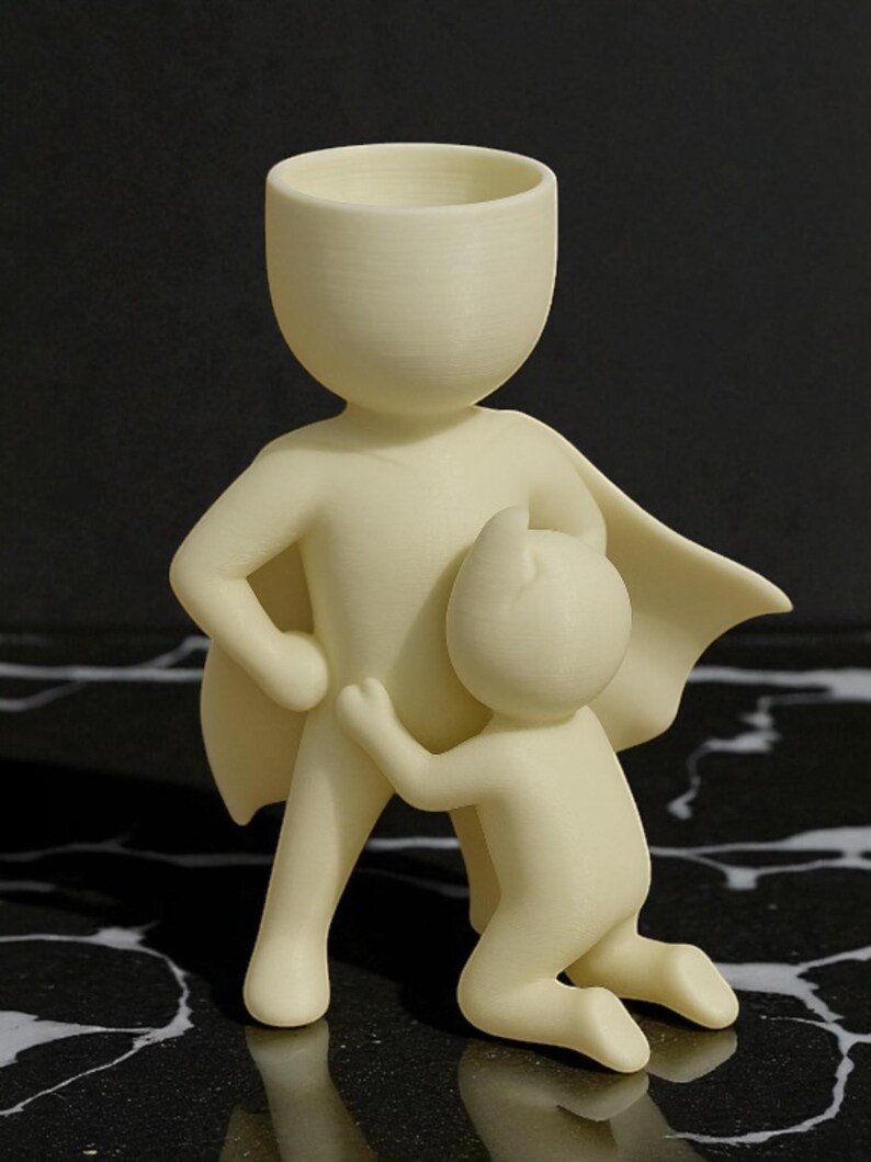Op de afbeelding: Een cr&egrave;mekleurig beeldje dat een ouderfiguur met een cape uitbeeldt, die een knielend kind omarmt. De figuren hebben eenvoudige, afgeronde vormen en staan tegen een donkere, patroonachtergrond. Het beeldhouwwerk is waarschijnlijk een decoratief item.