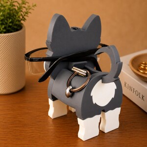 May include: A gray and white dog-shaped eyeglasses holder with black-framed glasses. The holder has a ring for holding keys or other small items. The dog-shaped holder is on a wooden surface.