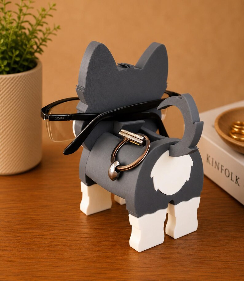 May include: A gray and white dog-shaped eyeglasses holder with black-framed glasses. The holder has a ring for holding keys or other small items. The dog-shaped holder is on a wooden surface.
