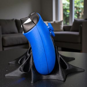 May include: A blue and black video game controller rests on a black, star-shaped stand. The controller has blue buttons and a black top. The stand is on a dark surface, with a blurred background of a living room.