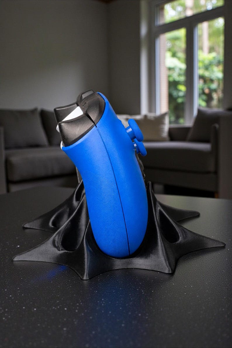 May include: A blue and black video game controller rests on a black, star-shaped stand. The controller has blue buttons and a black top. The stand is on a dark surface, with a blurred background of a living room.