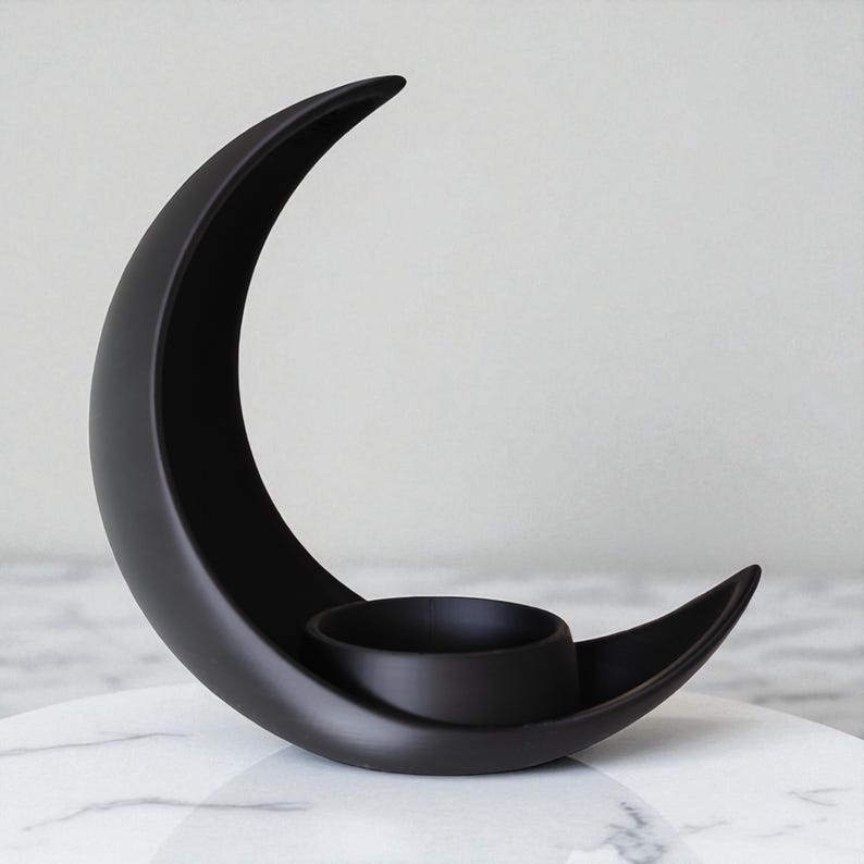 May include: A black ceramic crescent moon-shaped candle holder. The holder features a circular indentation for a candle and is set on a white marble surface. The design is minimalist and modern, suitable for home decor.