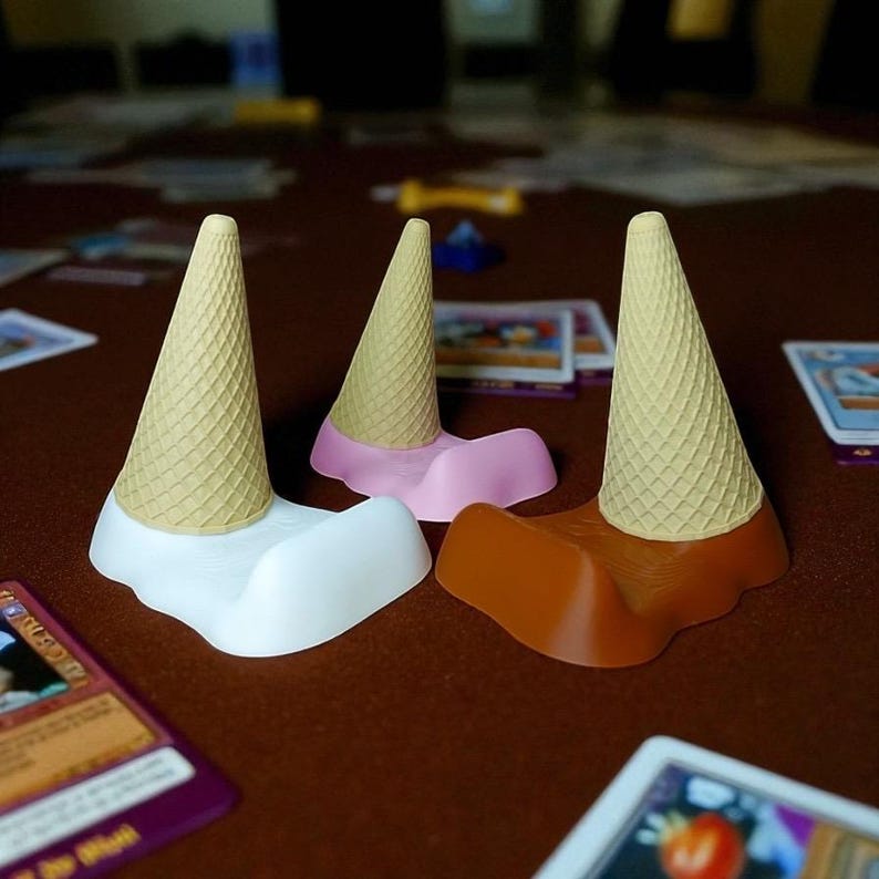 May include: Three colorful ice cream cone holders in white, pink, and brown. Each holder has a textured cone shape. The holders are on a brown surface with game cards in the background.