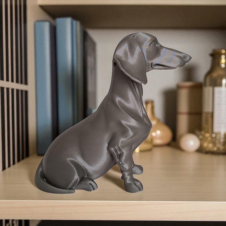May include: A gray dachshund figurine sitting on a wooden shelf. The dog statue is in a seated position, with its head turned to the right. The background includes books and decorative bottles.