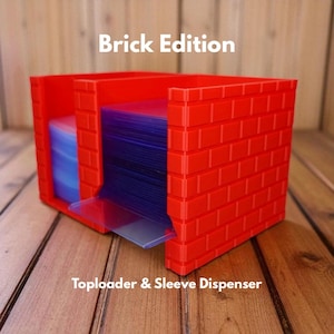 May include: A red plastic toploader and sleeve dispenser with a brick design. The dispenser has a clear plastic tray and holds blue and clear card sleeves. The text "Brick Edition" and "Toploader & Sleeve Dispenser" are visible.
