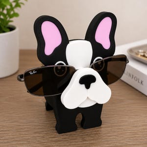 May include: A black, white, and pink bulldog-shaped eyeglass holder. The holder has a white snout, pink inner ears, and is wearing a pair of Ray-Ban sunglasses. The holder is made of a solid material and is sitting on a wooden surface.