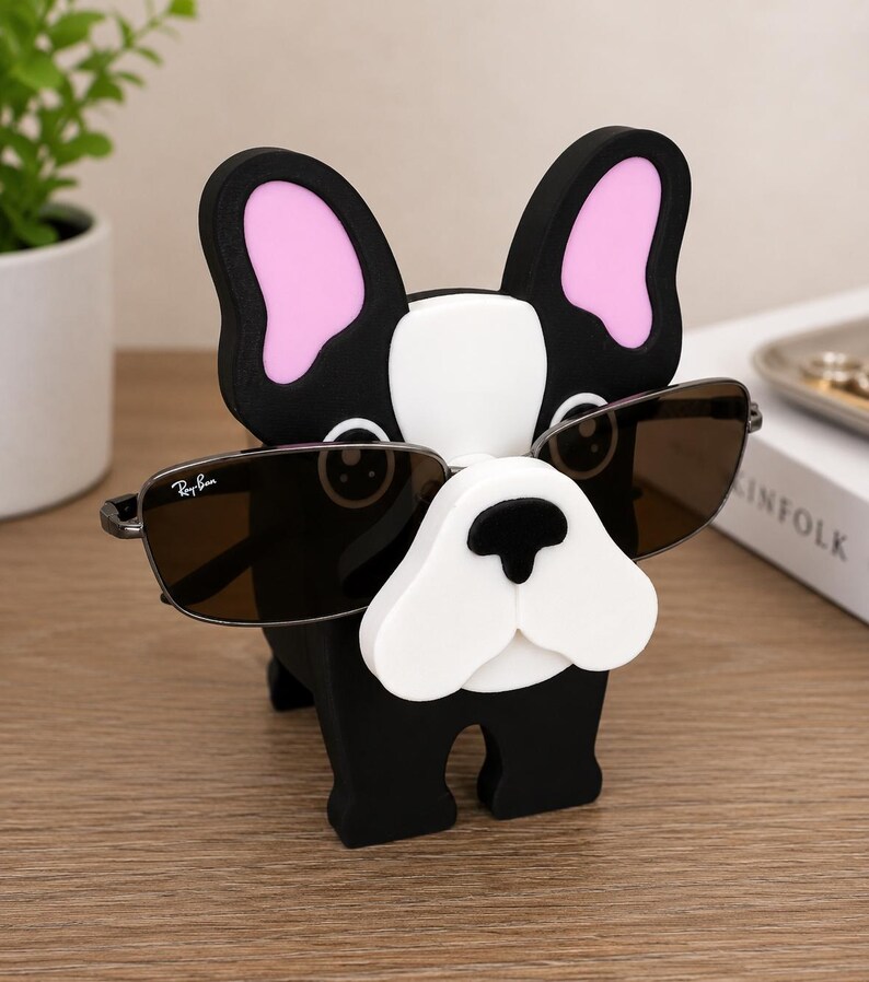 May include: A black, white, and pink bulldog-shaped eyeglass holder. The holder has a white snout, pink inner ears, and is wearing a pair of Ray-Ban sunglasses. The holder is made of a solid material and is sitting on a wooden surface.