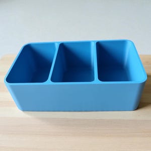Puede incluir: Un organizador de pl&aacute;stico azul claro con tres compartimentos de igual tama&ntilde;o. El contenedor rectangular se encuentra sobre una superficie de madera clara. El organizador est&aacute; dise&ntilde;ado para guardar y separar varios art&iacute;culos.