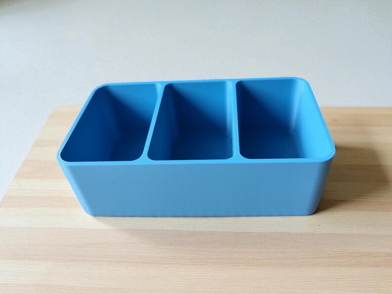 Puede incluir: Un organizador de pl&aacute;stico azul claro con tres compartimentos de igual tama&ntilde;o. El contenedor rectangular se encuentra sobre una superficie de madera clara. El organizador est&aacute; dise&ntilde;ado para guardar y separar varios art&iacute;culos.
