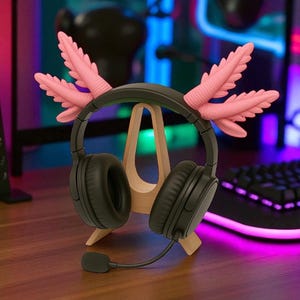Puede incluir: Auriculares negros de diadema con micr&oacute;fono, sobre un soporte de madera. Los auriculares tienen detalles rosas con forma de plumas. El soporte est&aacute; sobre una superficie de madera, con un teclado y fondo colorido.