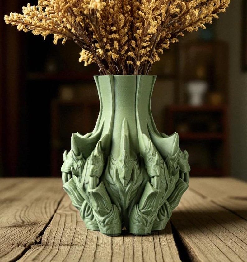 May include: A sage green decorative vase with a unique, textured design, holding dried, golden-brown flowers. The vase has an intricate, sculpted pattern and sits on a wooden surface, creating a natural aesthetic.