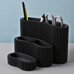 May include: A set of four black desk organizers with a ribbed design. The organizers vary in size and shape, with one holding pens, pencils, and scissors. The largest organizer is approximately 8 inches tall.