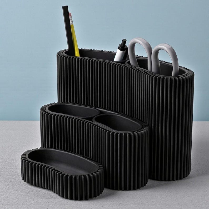 May include: A set of four black desk organizers with a ribbed design. The organizers vary in size and shape, with one holding pens, pencils, and scissors. The largest organizer is approximately 8 inches tall.