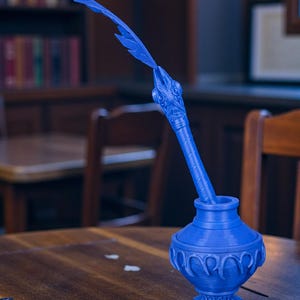 May include: A blue inkwell with a decorative feather pen. The inkwell has a detailed, ornate design and a round base. The feather pen is long and curved, with a detailed head. The inkwell and pen are made of a solid blue material.