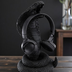 Op de afbeelding: Een zwarte koptelefoonstandaard in de vorm van een cobra, met een getextureerd oppervlak, die een zwarte JBL koptelefoon vasthoudt. De opgerolde standaard biedt een unieke presentatie voor audio-accessoires.