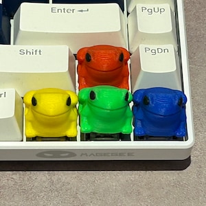 May include: Four frog-shaped keycaps in different colours: red, yellow, green, and blue. The keycaps are arranged in a row on a white keyboard.