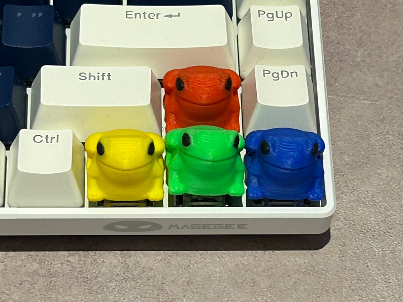 May include: Four frog-shaped keycaps in different colours: red, yellow, green, and blue. The keycaps are arranged in a row on a white keyboard.