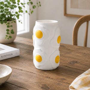 May include: White ceramic vase with a repeating pattern of yellow and white fried egg designs. The vase is cylindrical and sits on a wooden table. A small bowl and a plant in a pot are also visible.
