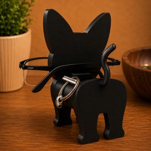 May include: A black cat-shaped eyeglasses holder with a tail and key ring. The holder is made of wood and has a pair of black-framed glasses resting on it. The cat is positioned on a wooden surface.