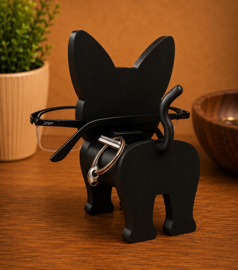 May include: A black cat-shaped eyeglasses holder with a tail and key ring. The holder is made of wood and has a pair of black-framed glasses resting on it. The cat is positioned on a wooden surface.