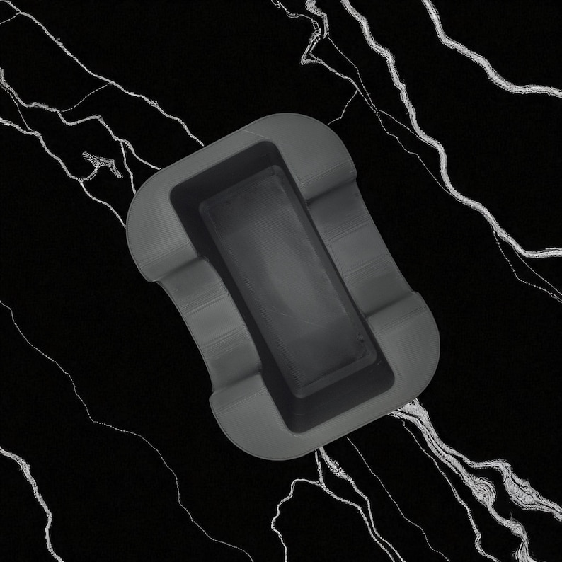 May include: A gray ashtray with a rectangular inner compartment and curved outer edges. The ashtray is made of a solid, matte material and sits on a black surface with white veining.
