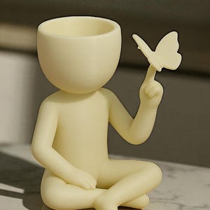 Puede incluir: Figura de color crema de una persona sentada con las piernas cruzadas, sosteniendo una mariposa en un dedo. La figura tiene una cabeza redondeada con una parte superior ahuecada, posiblemente para una peque&ntilde;a planta o decoraci&oacute;n. La mariposa tambi&eacute;n es de color crema.