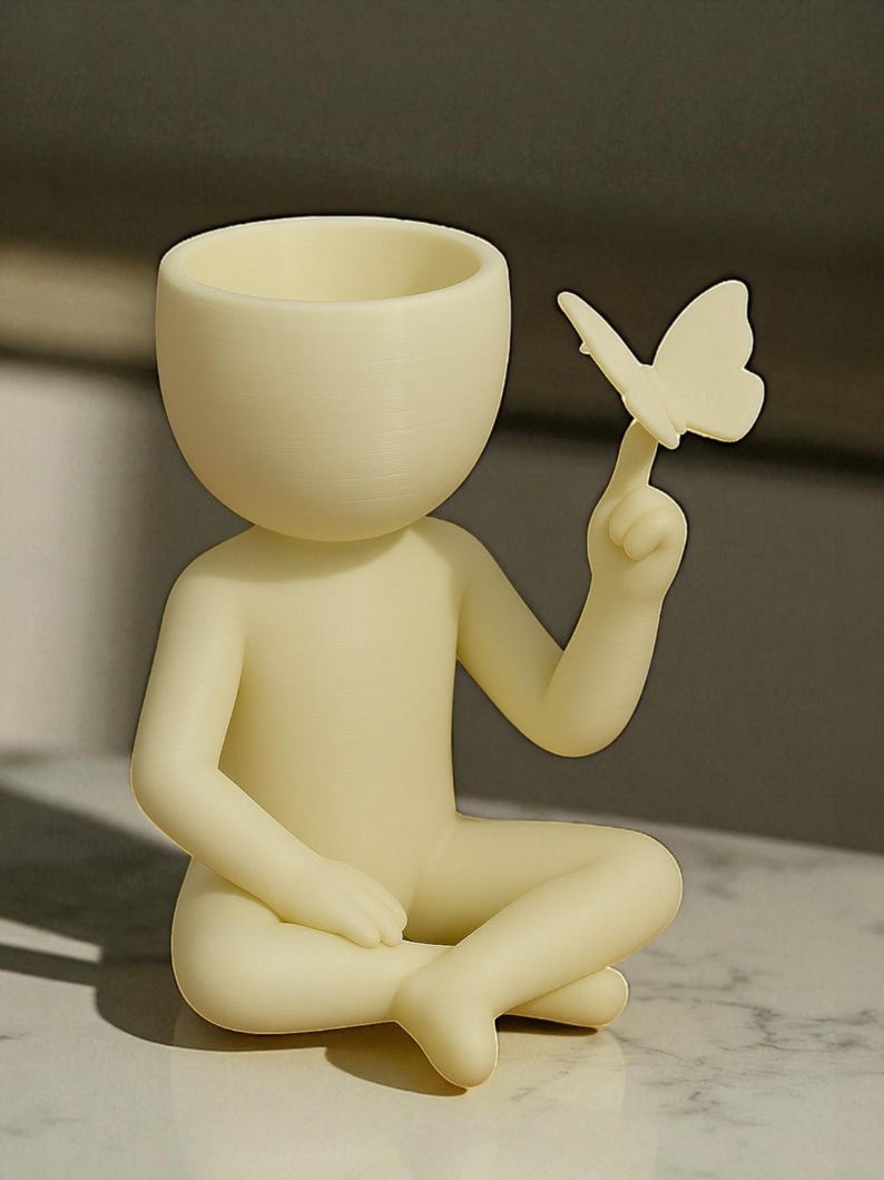Puede incluir: Figura de color crema de una persona sentada con las piernas cruzadas, sosteniendo una mariposa en un dedo. La figura tiene una cabeza redondeada con una parte superior ahuecada, posiblemente para una peque&ntilde;a planta o decoraci&oacute;n. La mariposa tambi&eacute;n es de color crema.