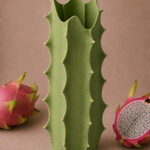 May include: A green cactus-shaped vase with a textured surface and small, light-colored protrusions. The vase is flanked by two dragon fruits, one whole and one cut open, revealing white flesh and black seeds. The background is a solid, neutral color.