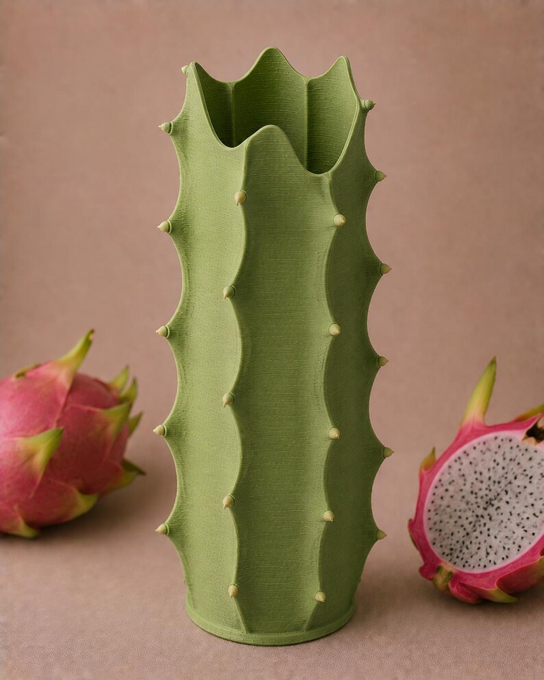 May include: A green cactus-shaped vase with a textured surface and small, light-colored protrusions. The vase is flanked by two dragon fruits, one whole and one cut open, revealing white flesh and black seeds. The background is a solid, neutral color.