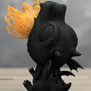 May include: A black, stylized dragon-like sculpture with a textured surface. The sculpture has a large head, small wings, and a tail. A cluster of orange and yellow flowers is placed on the dragon's head. The sculpture is on a wooden surface.