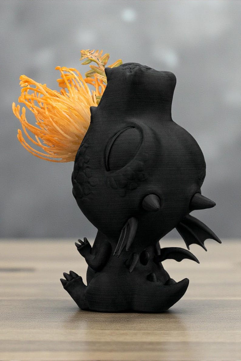 May include: A black, stylized dragon-like sculpture with a textured surface. The sculpture has a large head, small wings, and a tail. A cluster of orange and yellow flowers is placed on the dragon's head. The sculpture is on a wooden surface.