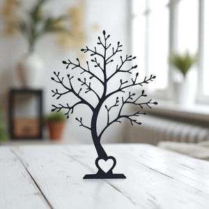 May include: A black metal tree-shaped jewelry holder with a heart-shaped cutout at the base. The tree has many branches with small, leaf-like details. The holder is displayed on a white wooden surface.