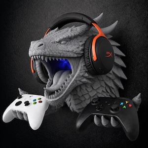 May include: A gray dragon head wall mount featuring a textured scale pattern and an open mouth with a blue light. The dragon wears black and orange over-ear headphones and holds a white game controller in one claw and a black one in the other.