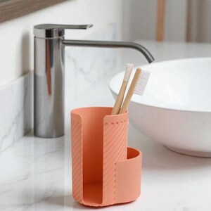 May include: A coral-colored toothbrush holder with two bamboo toothbrushes. The holder has a textured, ribbed design and a curved side. The bathroom accessory sits on a white marble countertop near a chrome faucet and a white sink.
