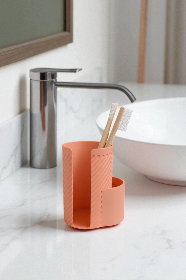 May include: A coral-colored toothbrush holder with two bamboo toothbrushes. The holder has a textured, ribbed design and a curved side. The bathroom accessory sits on a white marble countertop near a chrome faucet and a white sink.