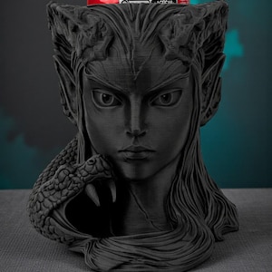 May include: A black, 3D-printed bust of a woman with long hair, pointed ears, and a snake wrapped around her. A red can sits in the bust's head. The bust is on a gray surface with a dark teal background.
